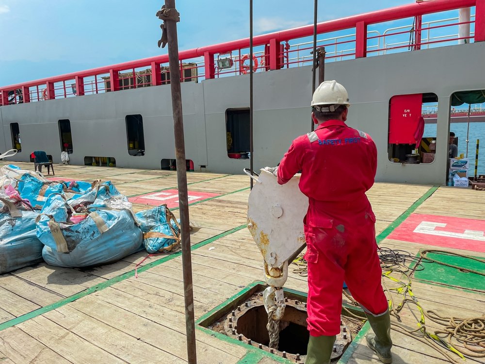 Deck worker in red coveralls guides a heavy-lift hook on a vessel deck near an open hatch, showing common maritime injuries hazards